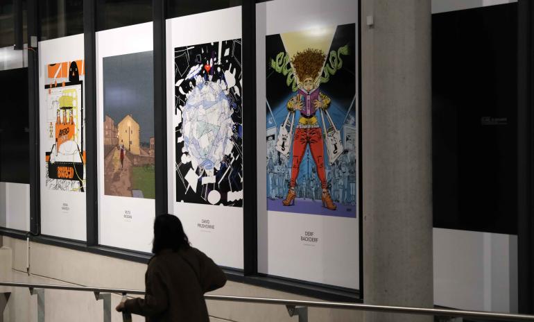 Photo d&#039;une femme avec une valise qui passe devant les panneaux de 4 illustrations de l&#039;expo &quot;50 ans, 50 regards&quot;.